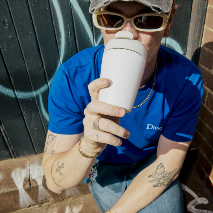 A person in a blue Dime t-shirt and sunglasses drinks from a KeepCup Commuter M 12oz in Bone White while sitting against a graffitied wall, showcasing the reusable cup's easy drinking lid.
