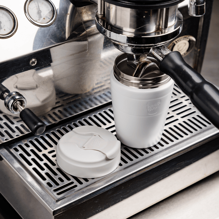 A KeepCup Commuter M 12oz in Bone White sits on the drip tray of a stainless steel espresso machine, with its matching white lid placed beside it, ready to catch a freshly pulled shot.
