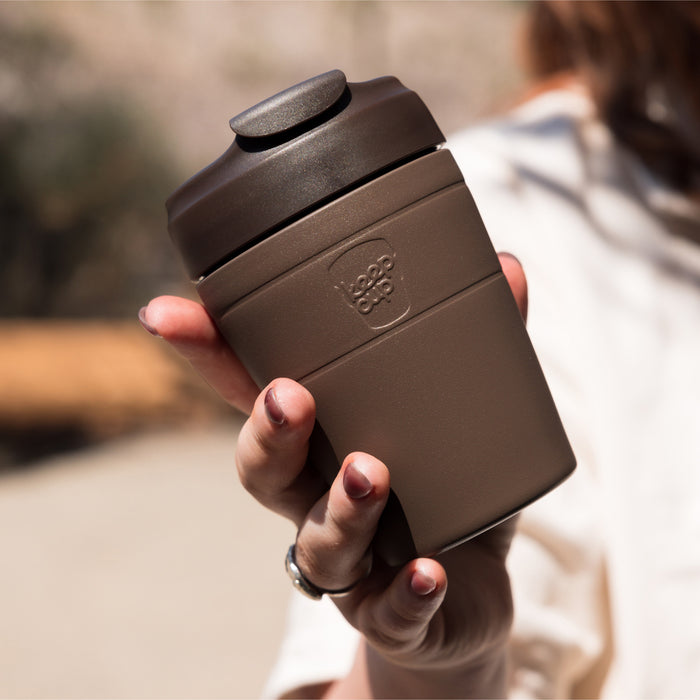 A hand holds a KeepCup Commuter M 12oz reusable coffee cup in Chocolate, showcasing its matte brown finish and embossed logo, with a soft-focus outdoor background.