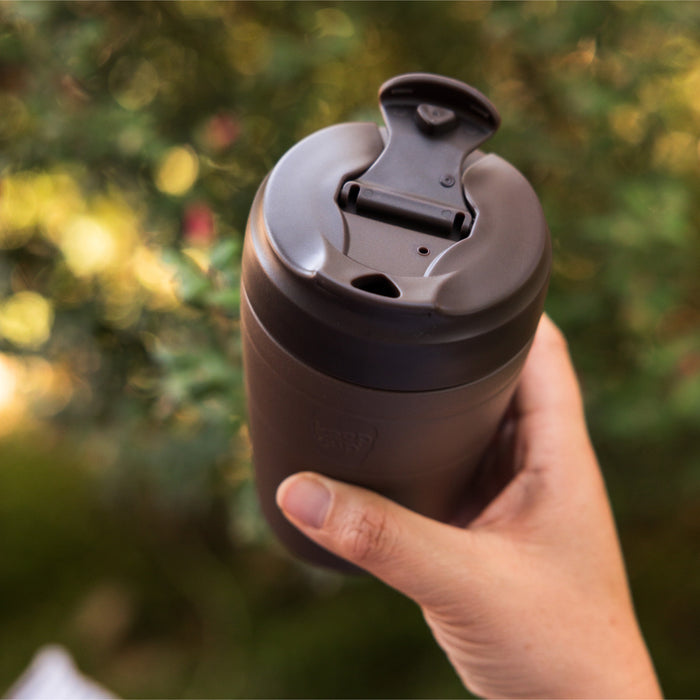 Hand holding a KeepCup Commuter M 12oz in Chocolate, showing the dark brown reusable cup with flip-top lid open, against a softly blurred outdoor background of green foliage.