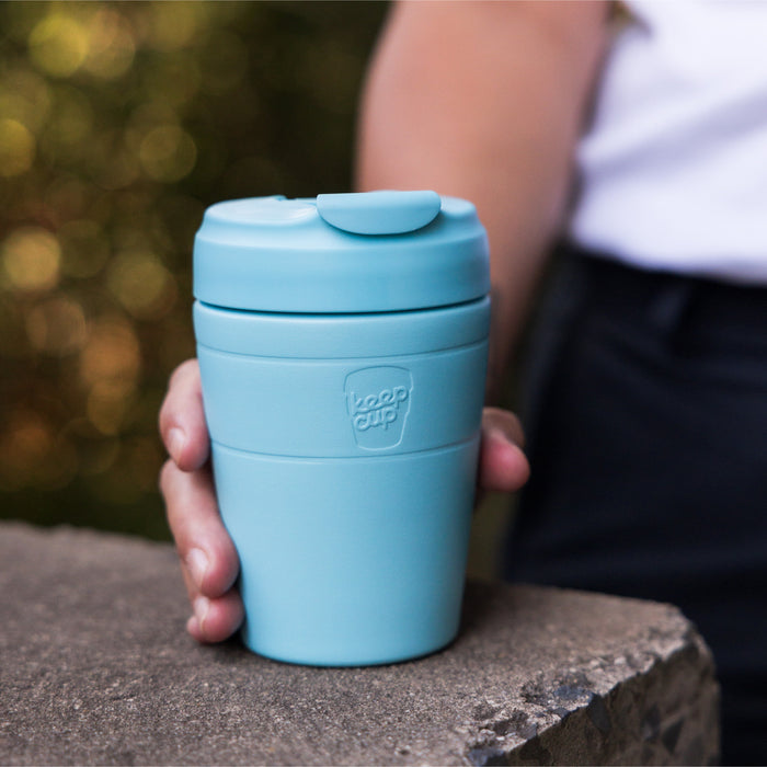 A hand rests on a stone ledge holding the KeepCup Commuter M 12oz in Cloud, a light blue reusable coffee cup with a flip-top lid, with soft bokeh greenery in the background.