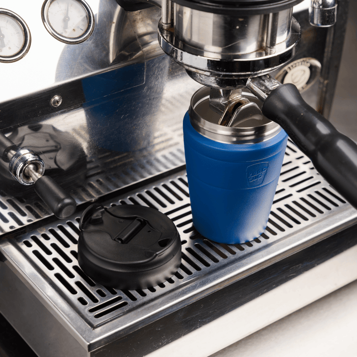 KeepCup Commuter M 12oz in Cobalt Black positioned under an espresso machine group head, with the black lid resting on the drip tray, demonstrating the cup's compatibility with café coffee machines.