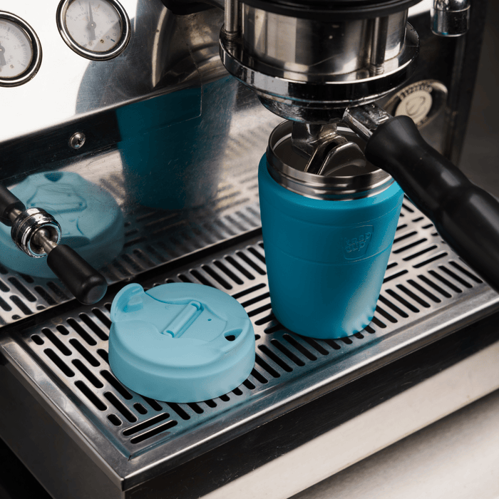 KeepCup Commuter M 12oz in Eden Cloud teal colour positioned under an espresso machine group head, with the light blue lid removed and placed on the stainless steel drip tray beside the cup.