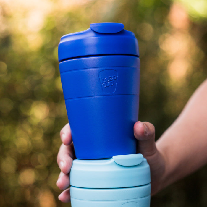 A hand holds the KeepCup Commuter M 12oz in Electric Blue, stacked on top of a light blue KeepCup, against a soft-focus outdoor background with natural greenery.