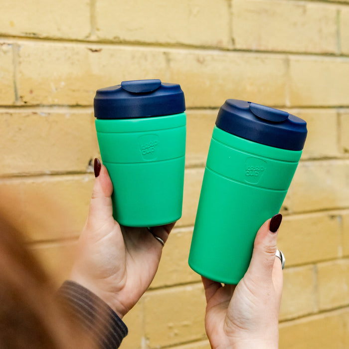Two hands with dark nail polish hold KeepCup Commuter M 12oz reusable cups in Emerald Ink, featuring bright green bodies and navy lids, against a yellow brick wall background.