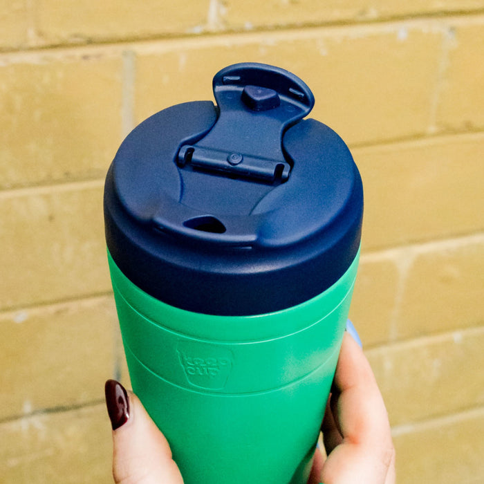 Hand holding a KeepCup Commuter M 12oz in Emerald Ink, featuring a bright green body and navy blue lid with flip-top opening, photographed against a yellow brick wall.