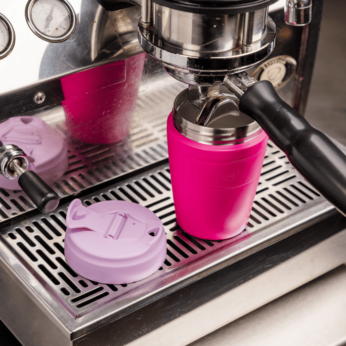 KeepCup Commuter M 12oz in Fuchsia Lilac positioned under an espresso machine group head, with the lilac lid removed and placed on the drip tray, demonstrating the cup's barista-friendly size for direct coffee extraction.