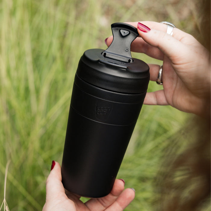 A woman's hands with dark red nail polish opening the flip-top lid of a black KeepCup Commuter L 16oz reusable coffee cup, with soft green grass in the background.