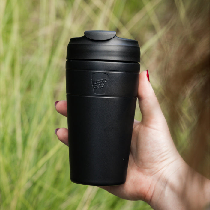 Hand holding a KeepCup Commuter L 16oz reusable coffee cup in Black, featuring a matte finish and matching black lid with sipper, set against a blurred outdoor background of tall grass.