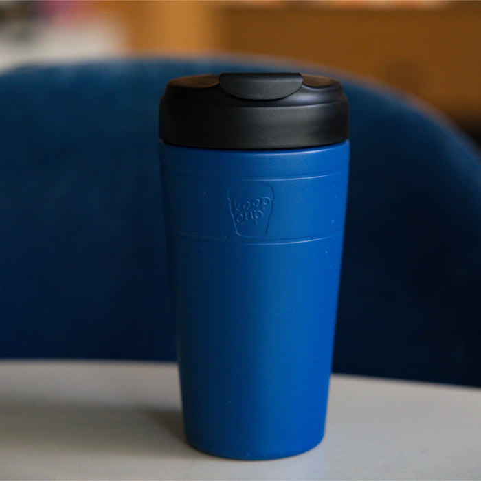 KeepCup Commuter L 16oz in Cobalt Black, featuring a blue body with black lid, sitting on a light grey surface with a blue chair in the background.