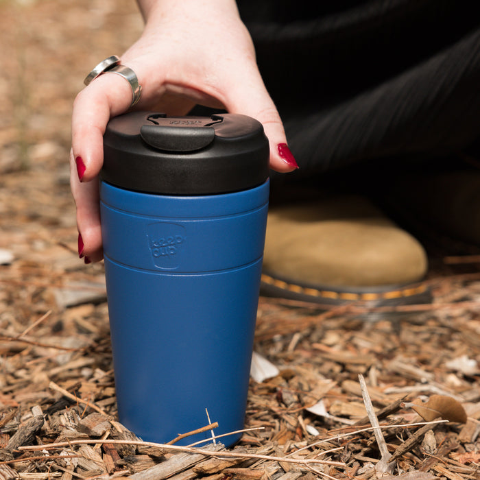 A hand with red nail polish reaches down to pick up a KeepCup Commuter L 16oz in Cobalt Black, resting on wood chip mulch outdoors, with the person's boots visible in the background.