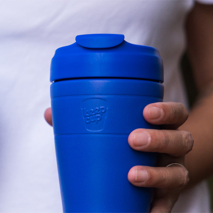 A hand holds the KeepCup Commuter L 16oz in Electric Blue, showing the vibrant blue reusable cup with its matching lid and sipper tab against a soft, blurred background.