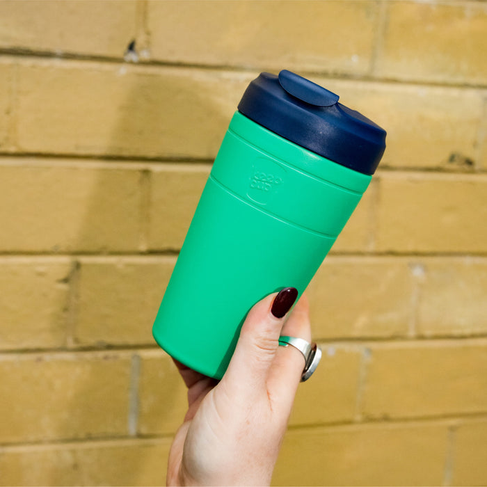 A hand with dark red nail polish and a silver ring holds a KeepCup Commuter L 16oz in Emerald Ink, featuring a bright green body and navy blue lid, against a yellow brick wall background.