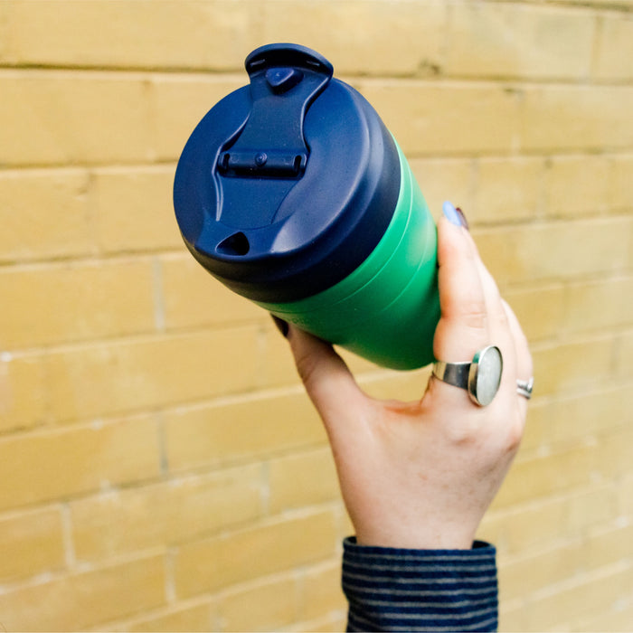 Hand holding a KeepCup Commuter L 16oz in Emerald Ink, featuring a green reusable cup body with navy blue lid, shown at an angle against a yellow brick wall.