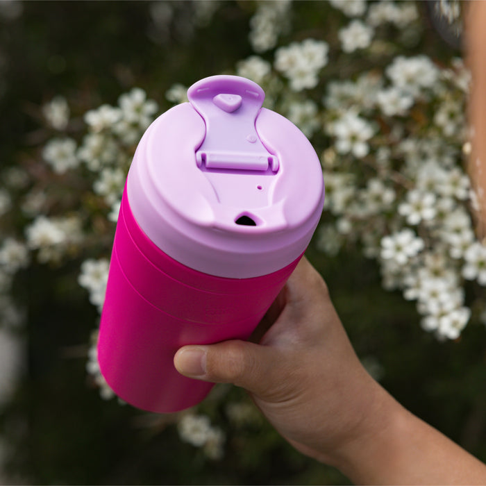 Hand holding a KeepCup Commuter L 16oz in Fuchsia Lilac, featuring a bright pink body with lilac lid shown at an angle to display the flip-top drinking spout, set against a soft-focus background of white blossoms.