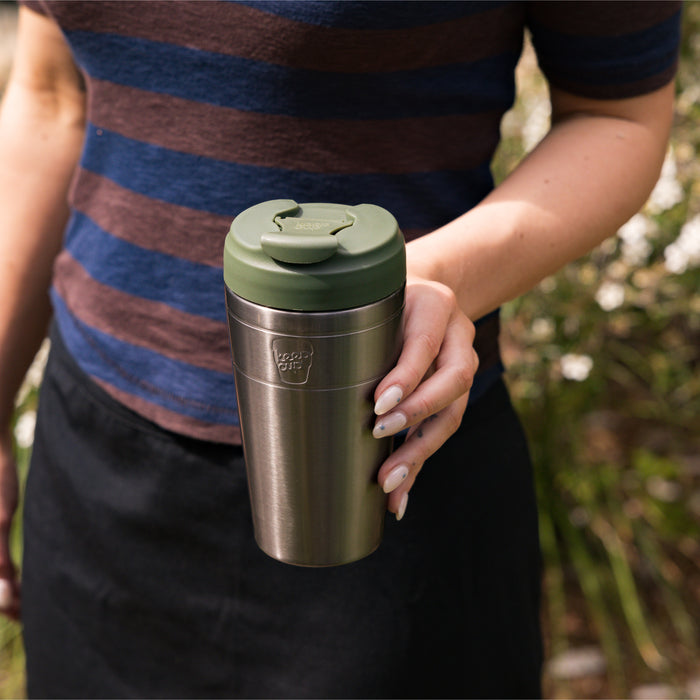 A hand with painted nails holds a KeepCup Commuter L 16oz in Nitro Pine, featuring a brushed stainless steel body and pine green lid, with a blurred outdoor background of greenery.