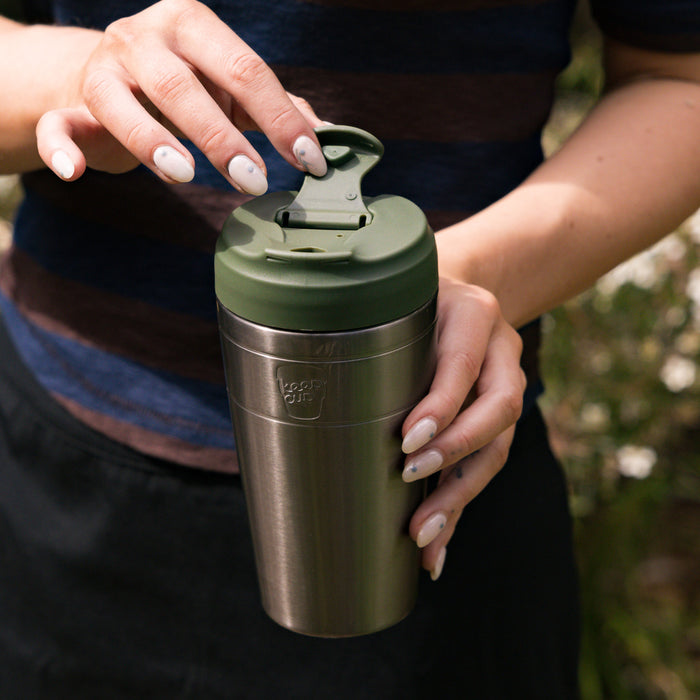 A person with light-coloured nail polish holds a KeepCup Commuter L 16oz in Nitro Pine, demonstrating the flip-top lid mechanism with their other hand, outdoors with greenery in the soft-focus background.