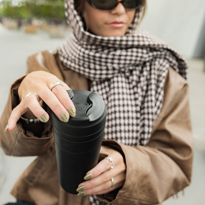 A woman wearing sunglasses, a brown jacket and checkered scarf holds a black KeepCup Commuter XL 20oz reusable cup, her olive-green manicured nails visible as she grips the lid in an outdoor urban setting.