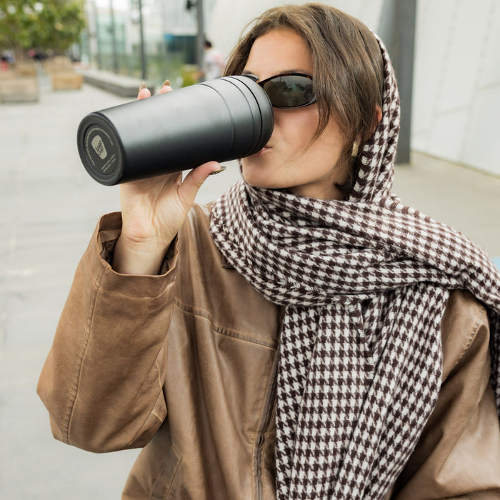 Woman wearing sunglasses and a houndstooth scarf drinks from a KeepCup Commuter XL 20oz in Black while outdoors, holding the reusable cup to her lips with one hand.