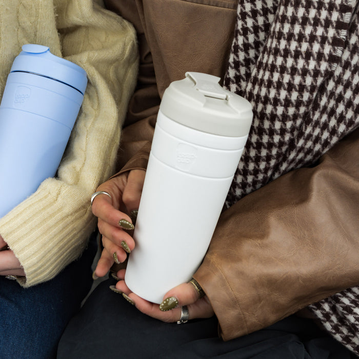 Two people sitting closely together, each holding a KeepCup Commuter XL 20oz reusable travel mug—one in Orchid and the other in the Bone White colour—showcasing the cups in a cosy, casual lifestyle setting with autumn knitwear and leather textures.