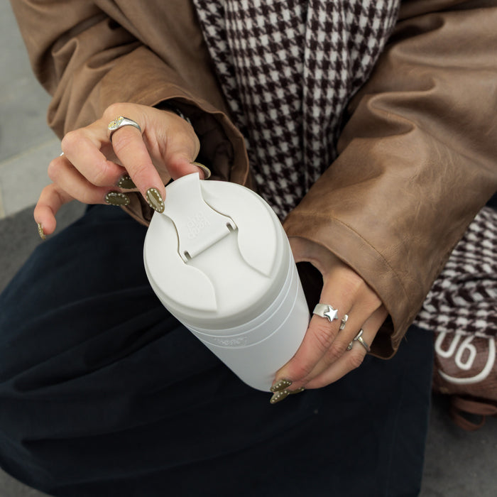 Hands with olive green nail art and silver rings hold a KeepCup Commuter XL in Bone White, opening the flip-top lid, styled with a brown leather jacket and houndstooth scarf.