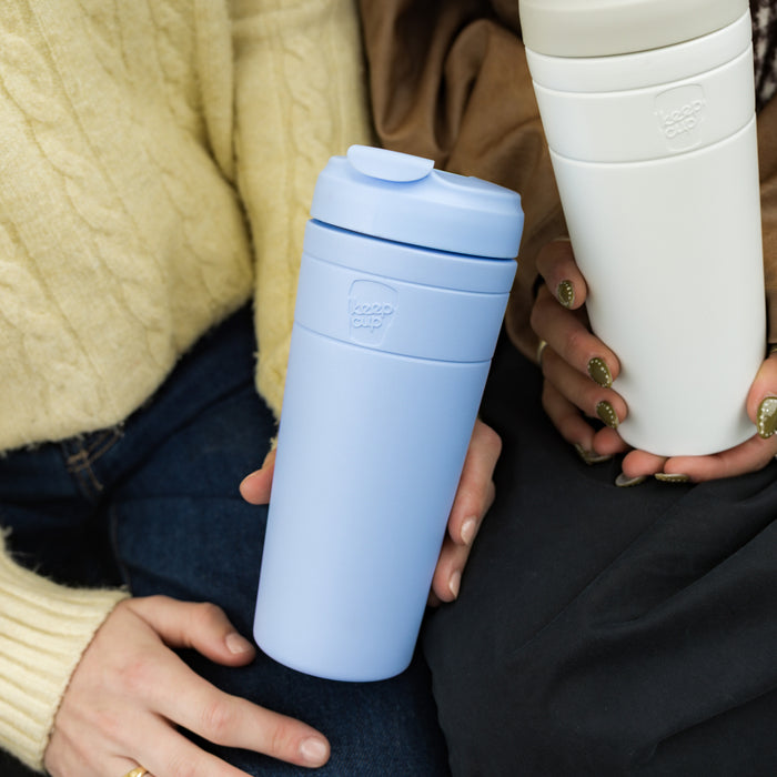 Two people sitting close together, each holding a KeepCup Commuter XL reusable cup—one in Orchid (light blue) and one in white—showcasing the cup's sleek design and comfortable grip.