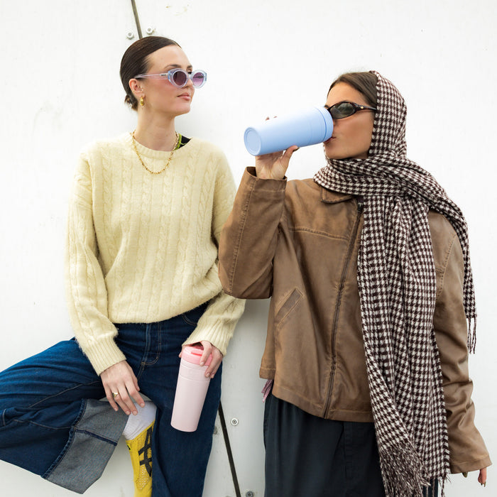 Two women in stylish autumn outfits lean against a white wall, one drinking from a KeepCup Commuter XL in White Pashmak while the other holds one in Orchid, showcasing the cups as fashionable everyday accessories.