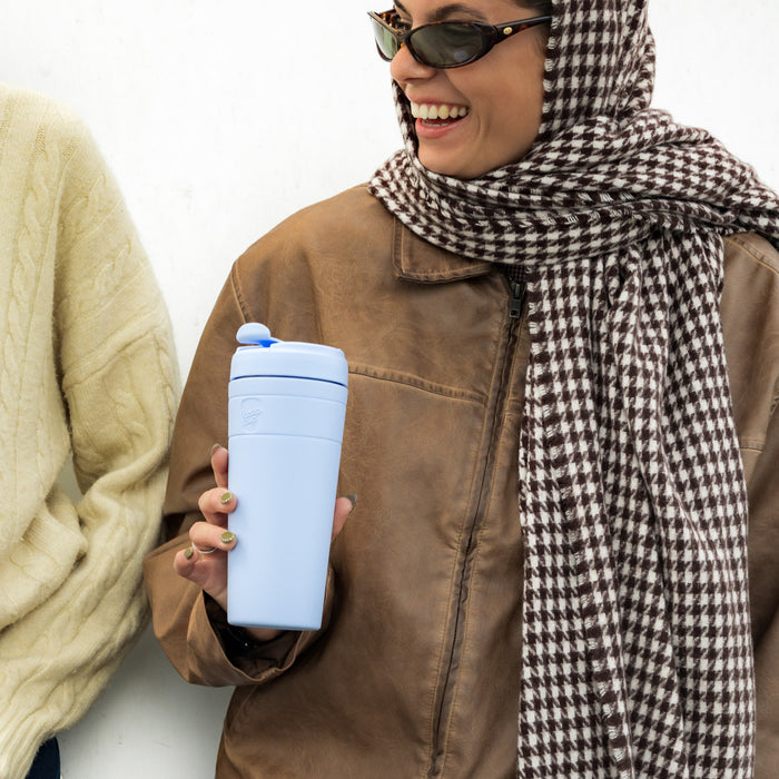 A smiling woman wearing sunglasses, a brown leather jacket and houndstooth scarf holds a KeepCup Commuter XL in Nitro Orchid, a light purple reusable cup with flip-top lid, while chatting with a friend outdoors.