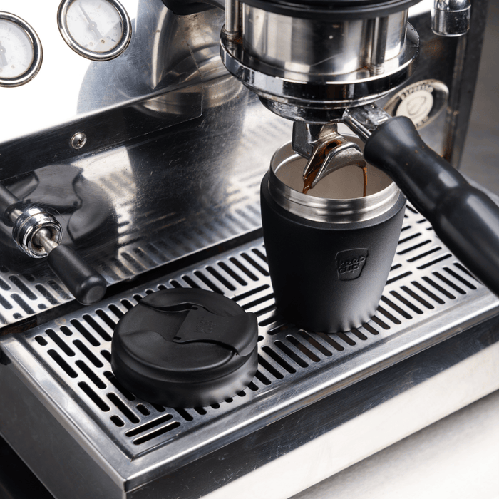 KeepCup Commuter S 8oz in Black positioned under an espresso machine group head with fresh coffee pouring into the stainless steel and silicone reusable cup, with the black lid resting on the drip tray beside it.