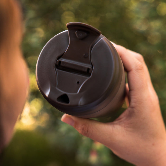 Hand holding a KeepCup Commuter S 8oz in Chocolate, viewed from above showing the dark brown flip-top lid with drinking spout, set against a soft-focus green outdoor background.