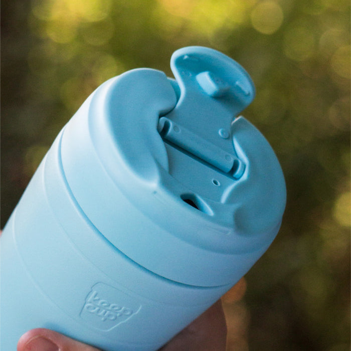 Close-up of a hand holding the KeepCup Commuter S 8oz in Cloud, showing the light blue lid with the sipper plug flipped open, set against a soft, blurred outdoor background.