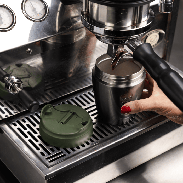 A hand with red nail polish holds a KeepCup Commuter S 8oz in Nitro Pine under an espresso machine portafilter, with the pine green lid placed on the metal drip tray beside it.