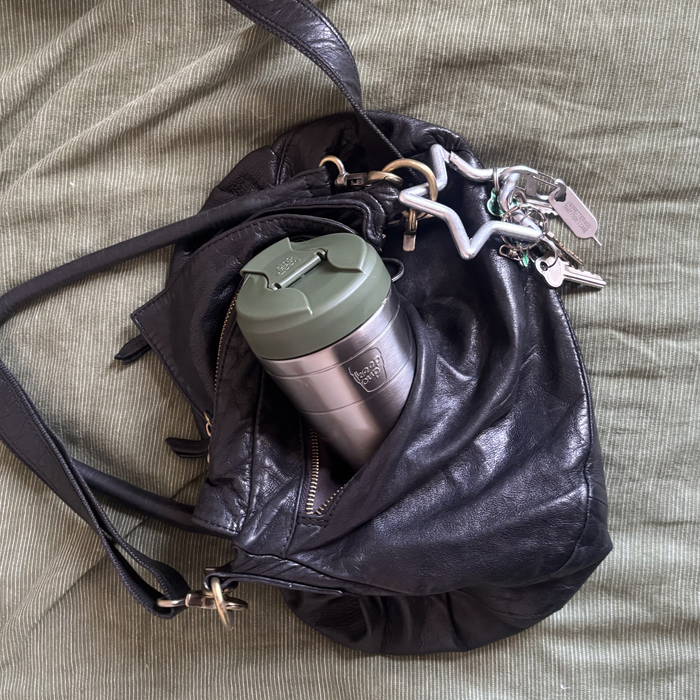 A black leather shoulder bag lying on a textured olive green fabric surface, with a silver insulated KeepCup travel mug and white keychain inserted into its side pocket.