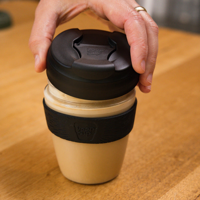 A hand reaches down to grasp the lid of a KeepCup Commuter Light M 12oz in Black, filled with a creamy coffee drink, sitting on a wooden table. The reusable cup features a beige body with a black silicone band and lid.
