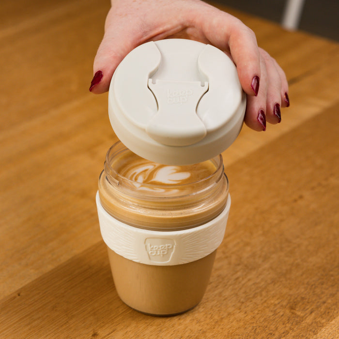 A hand with dark red nail polish lifts the white lid off a KeepCup Commuter Light M 12oz in Bone, revealing a latte with leaf-shaped latte art inside the glass cup, set on a wooden table.