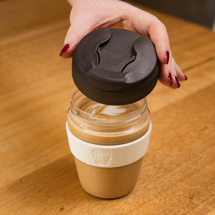 A hand with dark red nail polish lifts the charcoal lid from a KeepCup Commuter Light M 12oz in Chocolate, revealing a latte with leaf-shaped milk art inside, set on a wooden table.