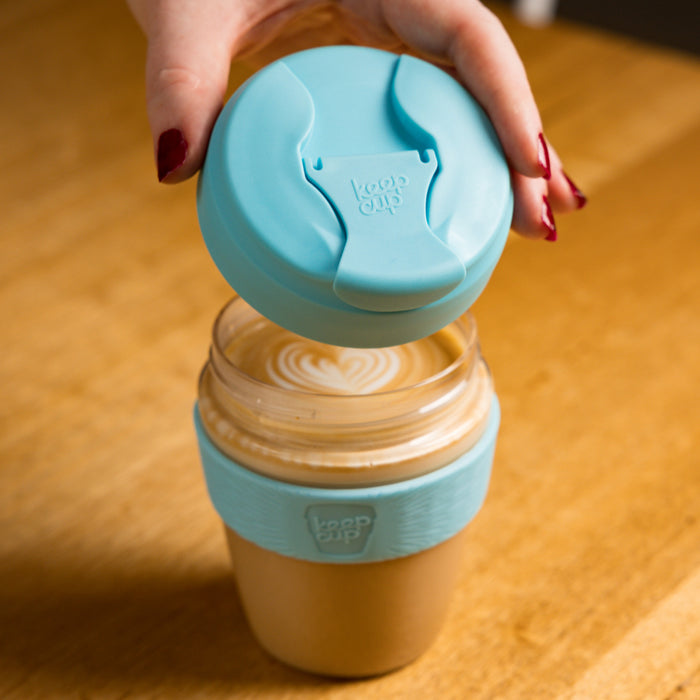 A hand with dark red nail polish lifts the light blue plastic lid from a KeepCup Commuter Light M 12oz in Cloud, revealing a latte with heart-shaped latte art inside the glass cup with matching blue band, set on a wooden table.