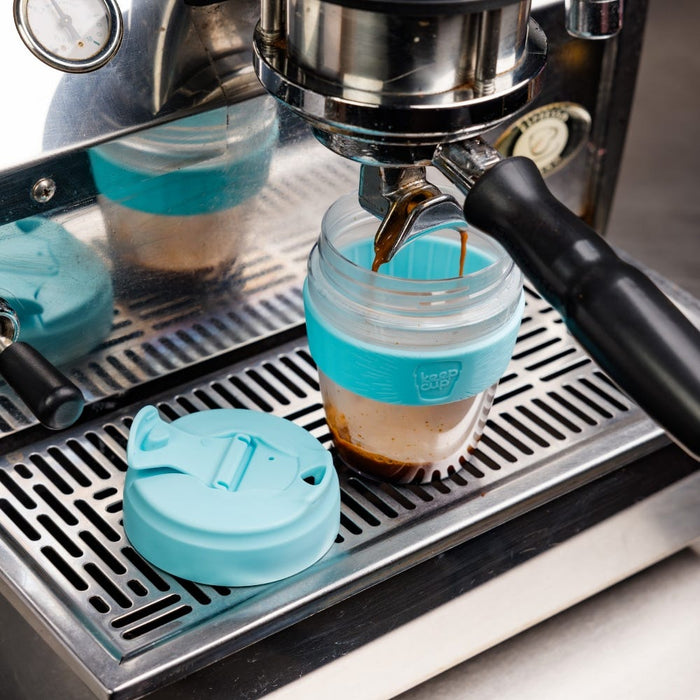 A KeepCup Commuter Light M 12oz in Cloud colour sits beneath an espresso machine portafilter with coffee streaming into it, the turquoise plastic lid resting beside it on the stainless steel drip tray.