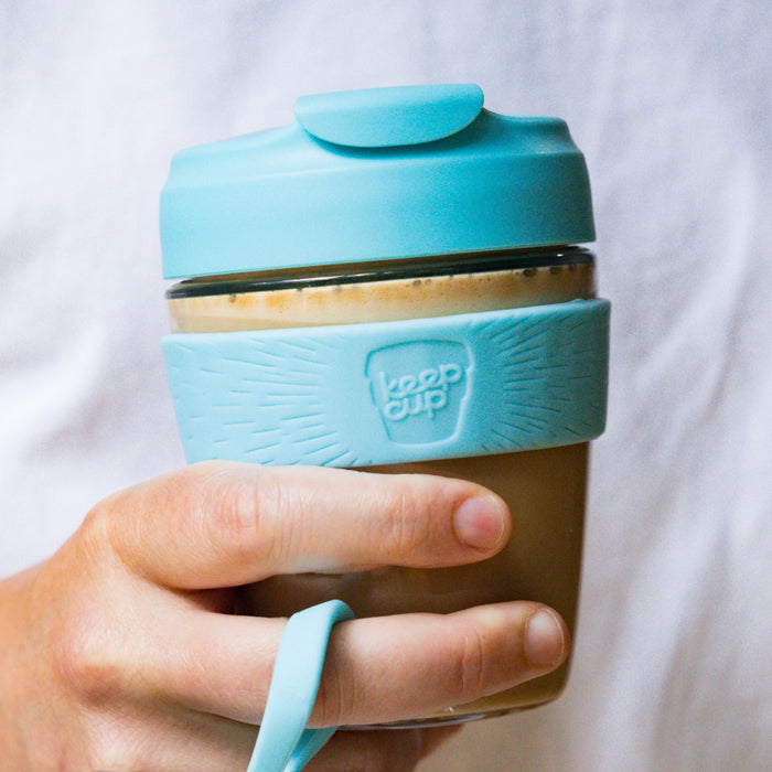 A hand holds a KeepCup Commuter Light M 12oz in Cloud, showing the light blue plastic lid and grip band with embossed KeepCup logo, wrapped around a glass cup filled with a latte.