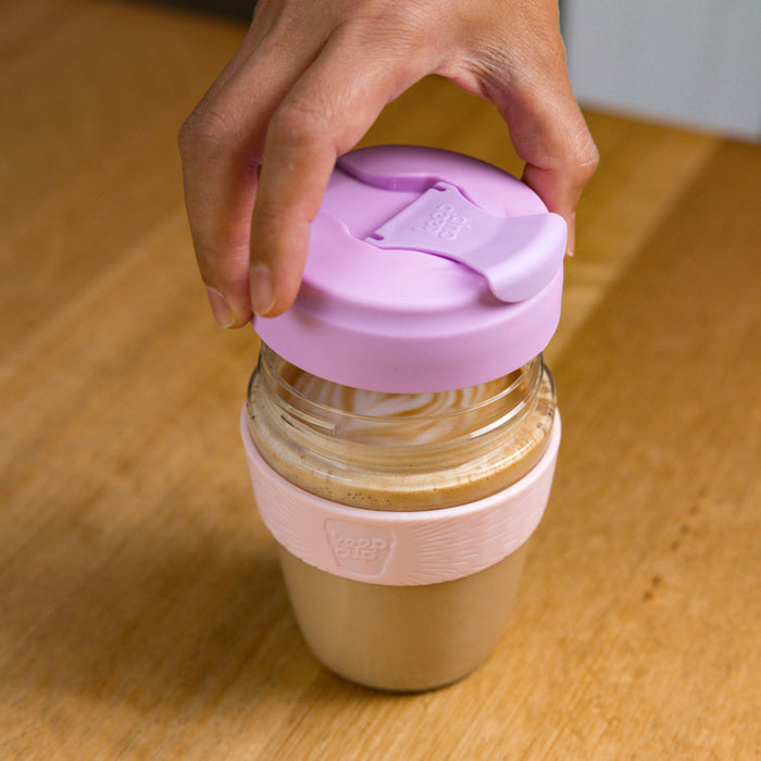 A hand lifting the lilac plastic lid off a KeepCup Commuter Light M 12oz filled with a latte, showing the glass body and pale pink band, placed on a wooden table.