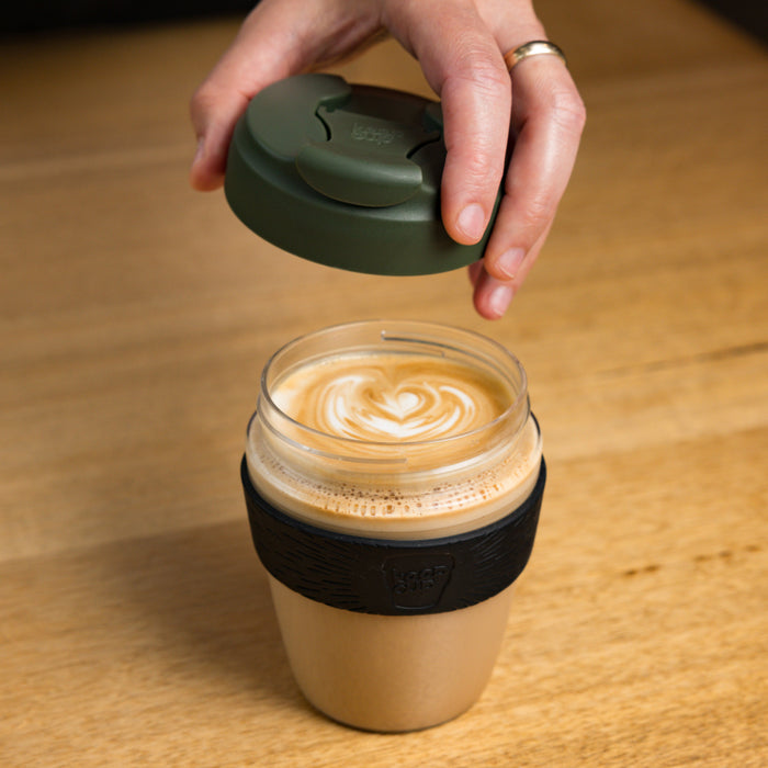 A hand lifts the Pine green lid from a KeepCup Commuter Light M 12oz reusable cup, revealing a latte with heart-shaped latte art inside the clear glass body with black silicone band, placed on a wooden table.