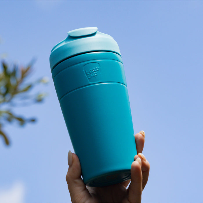 Hand holding a KeepCup Commuter L 16oz in Eden Cloud colour raised against a clear blue sky, showcasing the teal reusable cup's sleek design with its signature sipper lid and subtle branding.