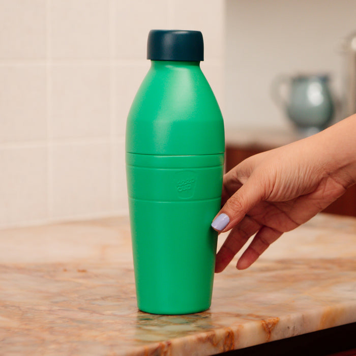 A hand with lavender nail polish reaches to grasp a KeepCup Deskside Warrior bottle in Emerald Cloud, featuring a vibrant green body and dark navy lid, positioned on a marble kitchen benchtop.