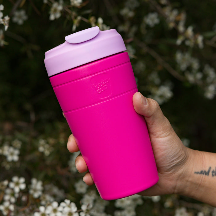 Hand holding a KeepCup Commuter L 16oz in Fuchsia Lilac, featuring a bright pink body with lilac lid, against a soft-focus background of white blossoms.