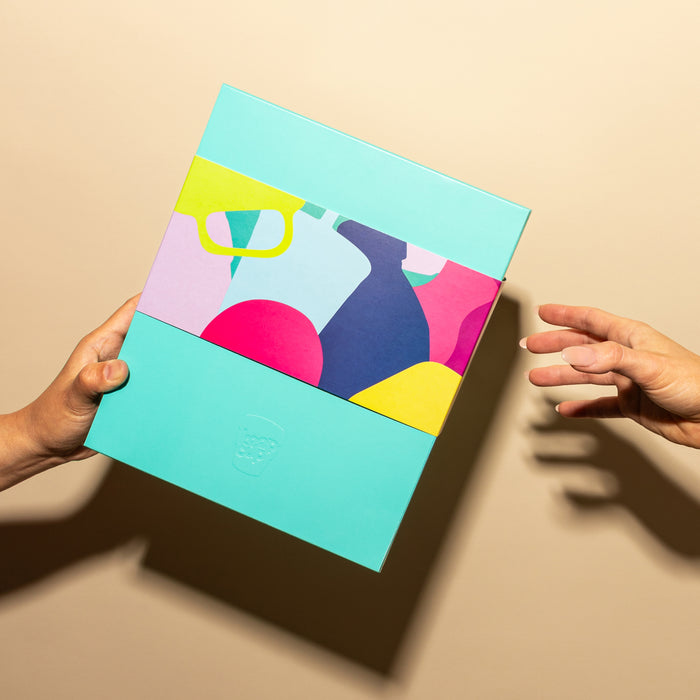 A hand extends a colourful KeepCup Deskside Warrior gift box in Cobalt Cloud towards another reaching hand, featuring vibrant abstract artwork with teal, pink, yellow and navy shapes on the sleeve, against a neutral beige background.