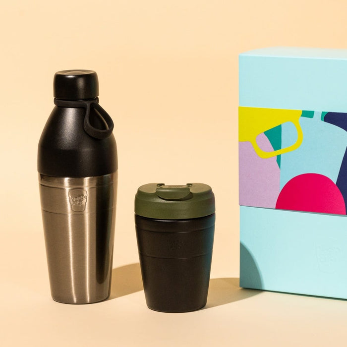 KeepCup Deskside Warrior gift set in Pine Black, featuring a stainless steel and black hybrid bottle with carry loop alongside a black reusable cup with olive green lid, displayed next to a colourful geometric gift box on a peach background.
