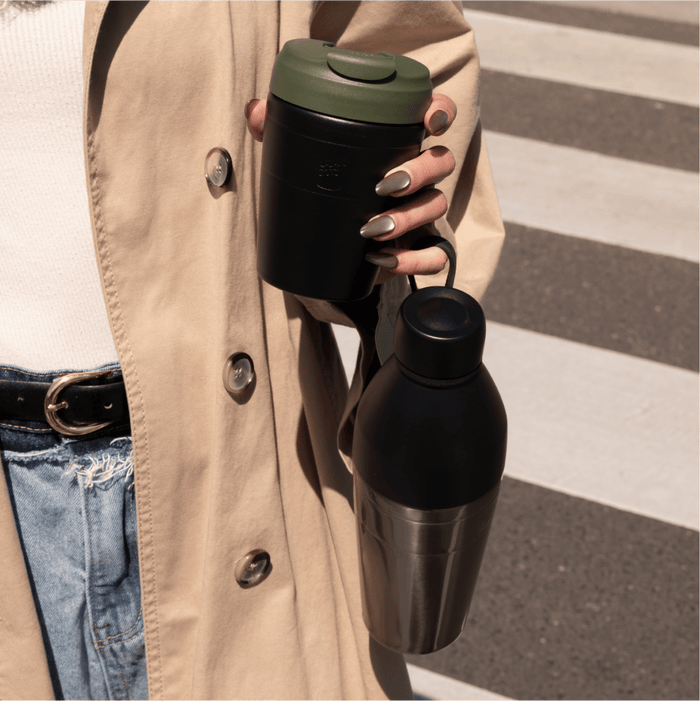 A woman in a tan trench coat holds the KeepCup Deskside Warrior set in Pine Black, featuring a black reusable coffee cup with olive green lid and a matching black stainless steel water bottle, while crossing at a pedestrian crossing.