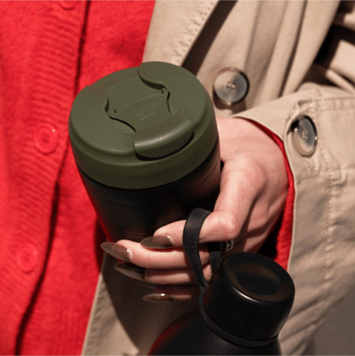 A hand holds the Deskside Warrior Pine Black KeepCup reusable coffee cup alongside a matching black water bottle with carry strap, against a backdrop of a red jumper and beige trench coat.