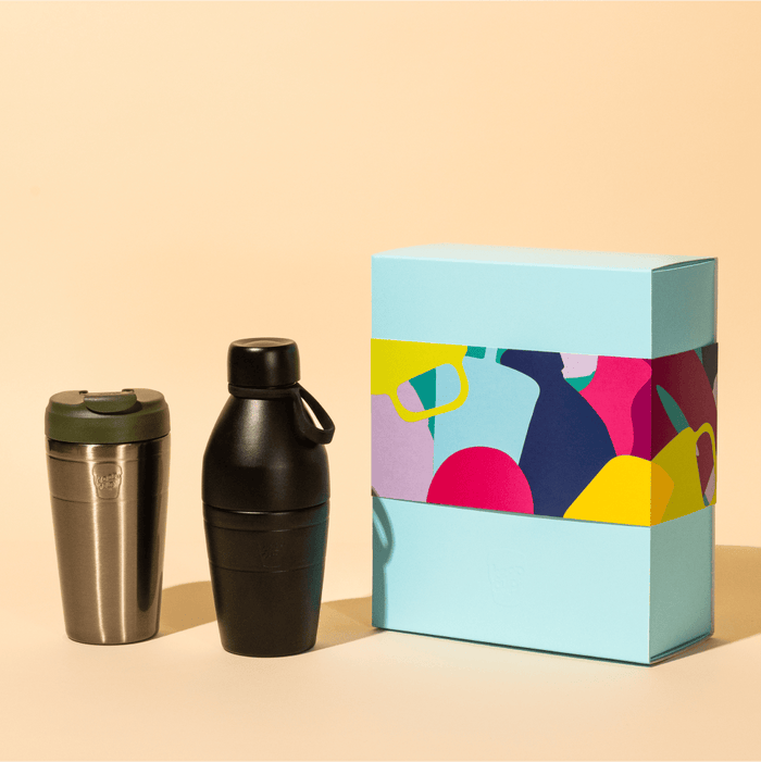 Deskside Warrior gift set featuring a stainless steel KeepCup with Pine green lid and a Black cocktail shaker-style bottle, displayed beside a light blue gift box with colourful abstract fruit artwork.