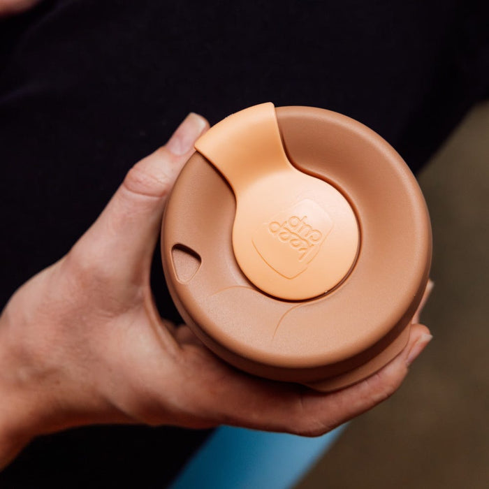Hand holding a KeepCup Brew Longplay M 12oz in Canna colour, viewed from above showing the warm terracotta-toned lid with lighter peach press-fit closure and embossed KeepCup logo.